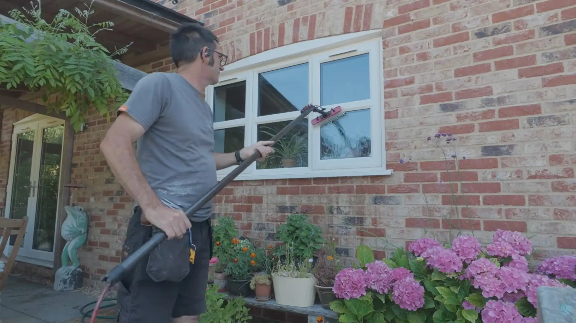 Technician using an extension pole to clean an exterior window near a garden bed